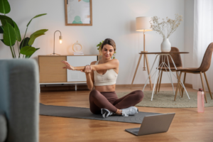 Mujer joven haciendo estiramientos en casa frente a su computadora portátil sobre un tapete de yoga.