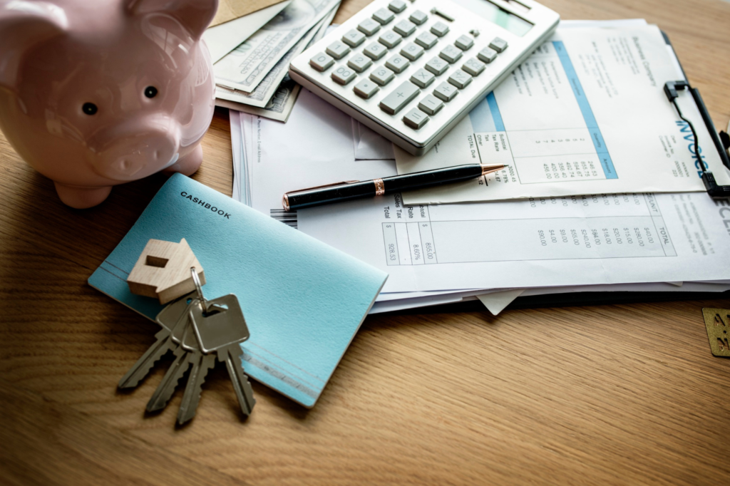 Mesa de madera con una alcancía de cerdito rosa, llaves con llavero de casa, libreta azul con la palabra “CASHBOOK”, una calculadora, bolígrafo negro, billetes de dólar y facturas. Representa el concepto de finanzas personales o administración del hogar.